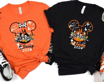 May include: An orange t-shirt and a black t-shirt, each featuring a graphic of Mickey Mouse ears filled with Disney characters in Halloween costumes, a castle, and the text Disney 2026. The shirts are displayed with black and orange sneakers.