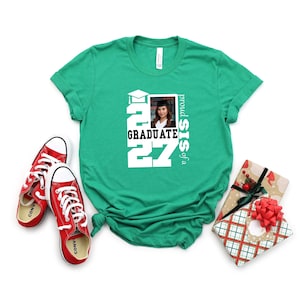 Op de afbeelding: Een felgroen T-shirt met korte mouwen en een witte opdruk met de tekst "proud SIS of a GRADUATE 2027" en een kleine rechthoekige foto van een persoon in een afstudeerhoed en toga, vergezeld door rode canvas sneakers.