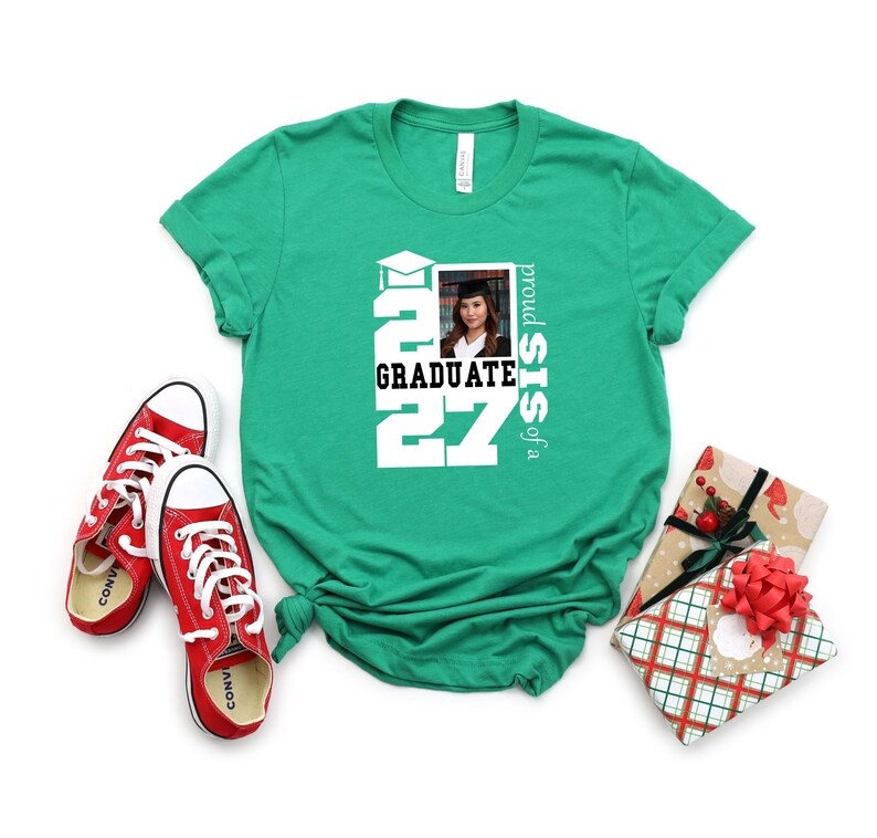 Op de afbeelding: Een felgroen T-shirt met korte mouwen en een witte opdruk met de tekst "proud SIS of a GRADUATE 2027" en een kleine rechthoekige foto van een persoon in een afstudeerhoed en toga, vergezeld door rode canvas sneakers.