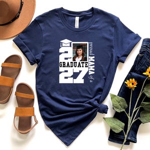 Op de afbeelding: Een marineblauw t-shirt met een wit grafisch ontwerp met de tekst "proud MAMA of a GRADUATE" en het jaar "2027" in grote blokletters, samen met een kleine vierkante foto van een persoon die een afstudeerhoed en toga draagt.