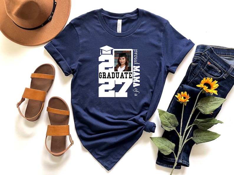 Op de afbeelding: Een marineblauw t-shirt met een wit grafisch ontwerp met de tekst "proud MAMA of a GRADUATE" en het jaar "2027" in grote blokletters, samen met een kleine vierkante foto van een persoon die een afstudeerhoed en toga draagt.