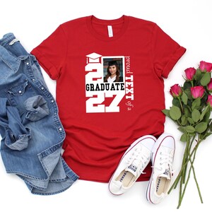 Op de afbeelding: Een rood T-shirt met korte mouwen en een witte afbeelding met de tekst 2027 GRADUATE, een icoon van een afstudeerhoed en een rechthoekig fotokader met een portret van een persoon in afstudeerkleding, met de tekst proud TEXT of a.