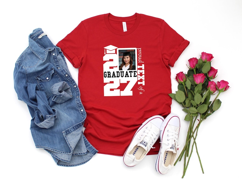 Op de afbeelding: Een rood T-shirt met korte mouwen en een witte afbeelding met de tekst 2027 GRADUATE, een icoon van een afstudeerhoed en een rechthoekig fotokader met een portret van een persoon in afstudeerkleding, met de tekst proud TEXT of a.