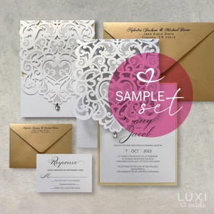May include: Elegant wedding invitation suite featuring laser-cut white lace designs, a sample set label, and gold envelopes. The invitation includes event details and a response card. A sparkling jewel adds a touch of glamour.