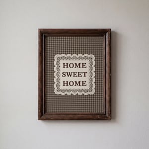 May include: A dark brown wooden frame containing a print with a brown and white gingham background. Centered on the print is a cream-coloured square with a decorative lace-style border, featuring the dark brown text HOME SWEET HOME.