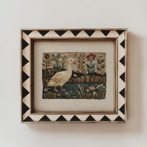 May include: A framed embroidery piece featuring a white goose standing in a landscape with colorful flowers and plants. The rectangular frame has a cream-colored border decorated with a repeating pattern of black triangles along the edges.