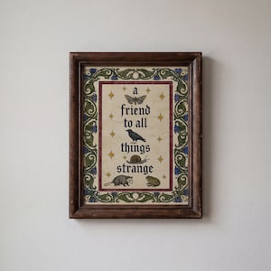 May include: A framed print featuring a dark brown wooden frame and a cream-coloured background with a green and blue floral border. The text reads 'a friend to all things strange' alongside illustrations of a moth, crow, snail, opossum, and frog.