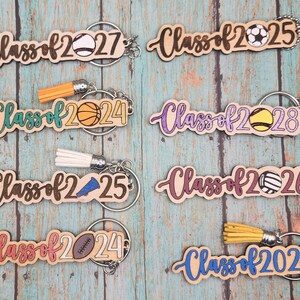 May include: Wooden keychains with the text "Class of" and a graduation year, featuring sports-themed designs like a baseball, basketball, soccer ball, volleyball, and football. Each keychain has a tassel.