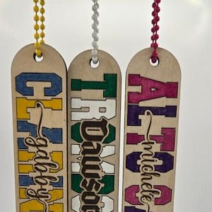 May include: Three wooden name tags with colorful lettering and beaded chains. The tags feature names in various fonts and colors: Gabby, Dawson, and Michele. Each tag is rectangular with a rounded top and a hole for the chain.