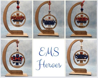 May include: Set of six wooden ornaments with blue and red star of life designs. Each ornament features a different phrase, including "Mommy", "Paramedic", "HER", "EMT", "Daddy", and "Medic 1".