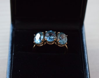 Puede incluir: Un anillo de oro con tres piedras preciosas ovaladas de color azul claro, presentado en una caja de joyería negra. La banda del anillo es dorada y las piedras están espaciadas uniformemente.