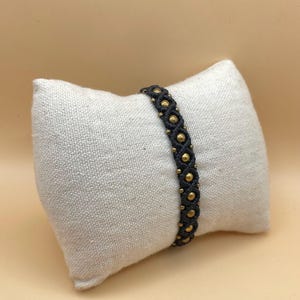 Op de afbeelding: Een donkergrijze macramé armband met goudkleurige kralen. De armband wordt getoond op een klein, off-white linnen kussen. De kralen zijn gelijkmatig verdeeld en geven het handgemaakte accessoire elegantie. Een stijlvol sieraad.