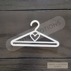 May include: White plastic hanger with a heart-shaped cutout in the top. The hanger is designed for small items like doll clothes or accessories.