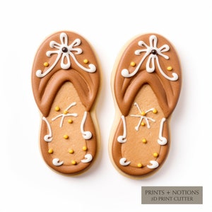 May include: Two brown gingerbread cookies decorated with white icing and yellow sprinkles, shaped like flip-flops. The cookies are on a white background.  PRINTS + NOTIONS 3D PRINT CUTTER
