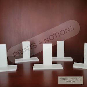 May include: Five white 3D printed display stands with a rectangular base and a vertical rectangular piece. The stands are arranged in a row on a brown surface. The text "PRINTS + NOTIONS 3D PRINT" is visible at the bottom of the image.