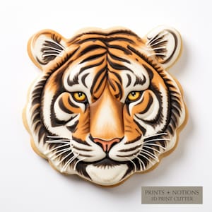 May include: A detailed cookie cutter in the shape of a tiger's face. The cutter is made of white plastic and has a detailed design of a tiger's face with black stripes, orange fur, and white whiskers. The text "PRINTS + NOTIONS 3D PRINT CUTTER" is printed on the bottom of the cutter.