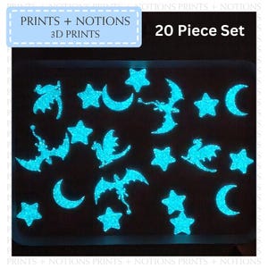 May include: A set of 20 glow-in-the-dark 3D printed stickers featuring stars, moons, and dragons. The stickers are arranged on a white tray and are glowing a bright blue color.
