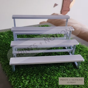 May include: A light gray, tiered display stand with four levels, each resembling a bench. The stand is set on a bed of artificial green grass. The text "PRINTS + NOTIONS 3D PRINT" is visible at the bottom.