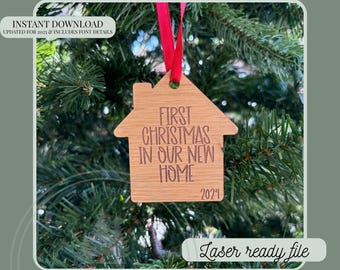 May include: A wooden house-shaped Christmas ornament with the text "First Christmas in Our New Home 2024". It hangs from a red ribbon against a backdrop of green pine needles. The ornament is a laser-ready file.