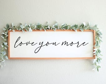 May include: A white wooden sign with the text "love you more" written in black cursive script. The sign is framed with a light brown wood frame and is decorated with a garland of green leaves.
