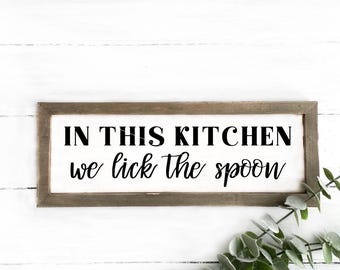 May include: A rustic wooden sign with the text "In this kitchen we lick the spoon" in black lettering. The sign is on a white wood background.