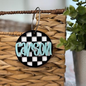 May include: A round, black and white checkered tag with the name "Carson" written in light blue. The tag is hanging from a woven basket.