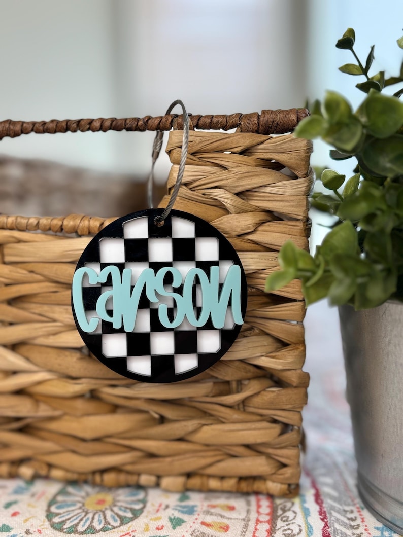 May include: A round, black and white checkered tag with the name "Carson" written in light blue. The tag is hanging from a woven basket.