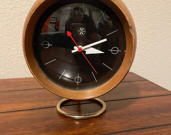 May include: A wooden clock with a black face and white hands. The clock is on a gold stand and has the word "Timelogic" printed on the face.