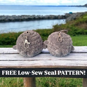 Könnte beinhalten: Zwei graue gehäkelte Robben-Plüschtiere mit Schlüsselanhängern. Die Plüschtiere sitzen auf einer Holzbank mit einem Gewässer im Hintergrund. FREE Low-Sew Seal Pattern! steht auf der Bank.