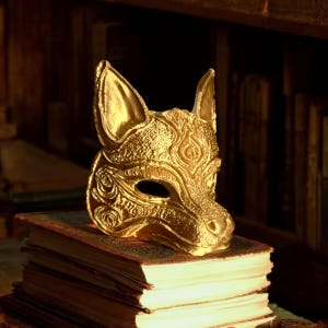 May include: A gold-coloured fox mask with detailed floral and swirl patterns, placed on a stack of antique books. The mask has a textured surface and a stylized design, suitable for costume or display.