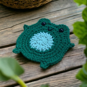 Könnte beinhalten: Handgefertigter grüner Häkel-Frosch-Untersetzer mit hellblauem Zentrum. Der Untersetzer hat zwei schwarze Knopfaugen und ein kleines aufgesticktes Lächeln. Der Frosch-Untersetzer liegt auf einer Holzoberfläche.