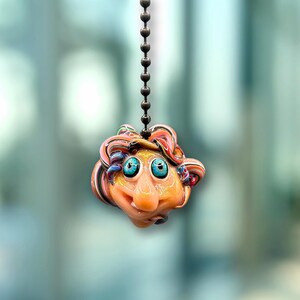 Pode incluir: Um pendente de rosto de vidro soprado à mão e caprichoso, pendurado numa corrente. O rosto é laranja com olhos azuis, um nariz grande e cabelo encaracolado colorido. A corrente é feita de metal escuro.