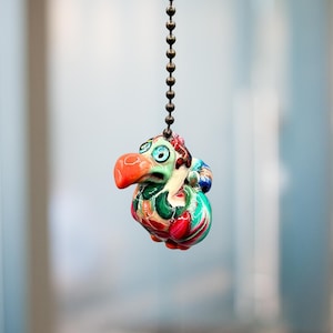 May include: A colorful, whimsical ceramic dodo bird light pull. The bird features a bright orange beak, blue eyes, and a patterned body with green, red, and white accents. It hangs from a bronze-colored chain.