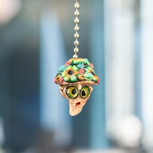 May include: A novelty mushroom-shaped ceiling fan pull. The mushroom cap is adorned with a vibrant floral design in green, blue, and red. The mushroom has large, surprised green eyes and a surprised expression. It is suspended from a silver chain.