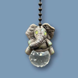 Puede incluir: Una figura decorativa de elefante con una base de bola de cristal, colgando de una cadena. El elefante es gris con acentos iridiscentes y ojos amarillos. La bola de cristal está facetada, reflejando la luz.