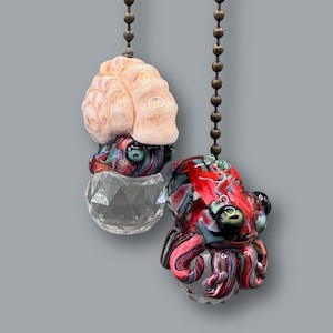 May include: Two decorative glass octopus-shaped ceiling fan pulls. One is light pink and the other is red, blue, and black. Both have a clear crystal base and hang from a bronze-coloured chain.