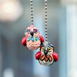 May include: Two decorative owl light pull chains. One is pink with red accents, the other is white with red accents. Both have detailed designs and hang from silver ball chains.