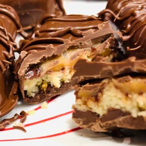 May include: Close-up of a chocolate bar with a caramel and coconut filling. The bar is broken in half, revealing the layers of filling. The chocolate is dark brown and shiny, and the caramel is a light golden brown. The coconut is white and fluffy.