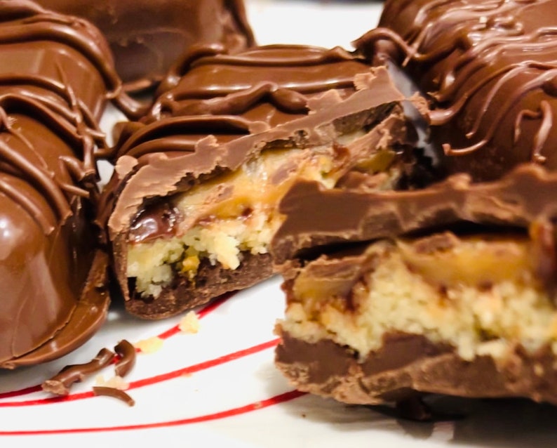 May include: Close-up of a chocolate bar with a caramel and coconut filling. The bar is broken in half, revealing the layers of filling. The chocolate is dark brown and shiny, and the caramel is a light golden brown. The coconut is white and fluffy.