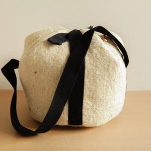 以下が含まれることがあります： 黒いストラップが付いた白い丸いフェルトバッグ。バッグは巾着式で、柔らかく質感のある素材で作られています。