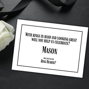Peut inclure: Une carte blanche avec du texte noir demandant "Avec des anneaux en main et en ayant fière allure, voulez-vous nous aider à célébrer ? Mason Acceptez-vous d'être notre porteur d'alliances ?"