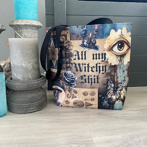 May include: A black tote bag with a colorful design featuring a witchy theme. The bag has a white background with a variety of images, including a large eye, a butterfly, and a floral design. The text "All my Witchy Shit" is printed in black on the bag.