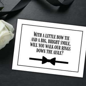 Peut inclure: Une carte blanche avec du texte noir qui dit "WITH A LITTLE BOW TIE AND A BIG, BRIGHT SMILE, WILL YOU WALK OUR RINGS DOWN THE AISLE?" Un nœud papillon noir est illustré sous le texte.