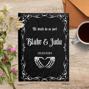 May include: Black wedding guest book with a white ornate frame, the text "Till death do us part", "Blake & Jada", and "04-23-2024" and a pair of white skeletal hands forming a heart.