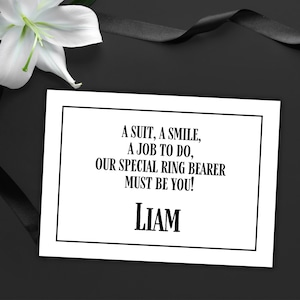 Peut inclure: Une carte blanche avec une bordure noire et le texte "A SUIT, A SMILE, A JOB TO DO, OUR SPECIAL RING BEARER MUST BE YOU!" et le nom "LIAM". Une fleur blanche et un ruban noir sont en arrière-plan.