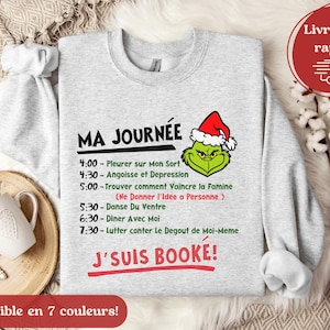 Peut inclure: Un sweat-shirt gris avec un personnage vert du Grinch portant un chapeau de Père Noël. Le texte sur le sweat-shirt indique "MA JOURNÉE" et liste un horaire quotidien humoristique. Le texte en bas indique "J'SUIS BOOKE!"