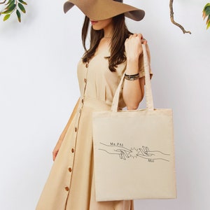 Puede incluir: Una bolsa de tela beige con un dibujo de línea negra de dos manos que se alcanzan. El texto "Ma PAL" y "Moi" está impreso en la bolsa.
