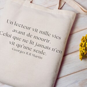 Può includere: Una borsa di tela bianca con una citazione di George R.R. Martin stampata su di essa. La citazione recita: "Un lettore vive mille vite prima di morire. Chi non legge ne vive solo una." La borsa è appoggiata su una superficie di legno con un mazzo di fiori gialli a destra.