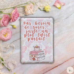 Puede incluir: Una funda para iPad rosa y blanca con una cita en francés que dice "Pas besoin de roses, juste un plot twist parfait." La cita está rodeada de corazones rosas. Una pila de libros con una vela encendida en la parte superior está en el centro de la funda.