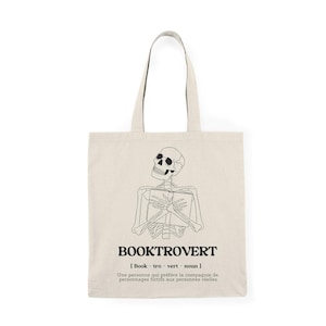 Peut inclure: Un sac fourre-tout beige avec un contour noir d'un squelette tenant un livre. Le sac porte le texte "BOOKTROVERT" en noir avec la définition "[Book - tro - vert - noun] Une personne qui préfère la compagnie de personnages fictifs aux personnes réelles."